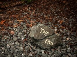 A rock with the words "THE END" painted on it, resting on gravel and surrounded by fallen leaves and twigs.