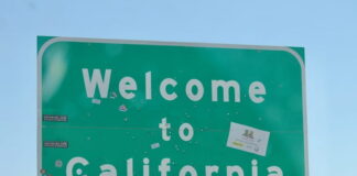 Green 'Welcome to California' road sign covered with various stickers against a clear blue sky.