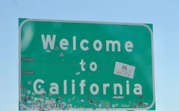 Green 'Welcome to California' road sign covered with various stickers against a clear blue sky.